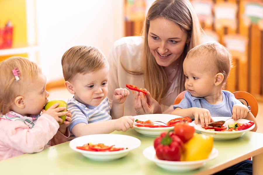 woman crouching next to babies eating at a small table